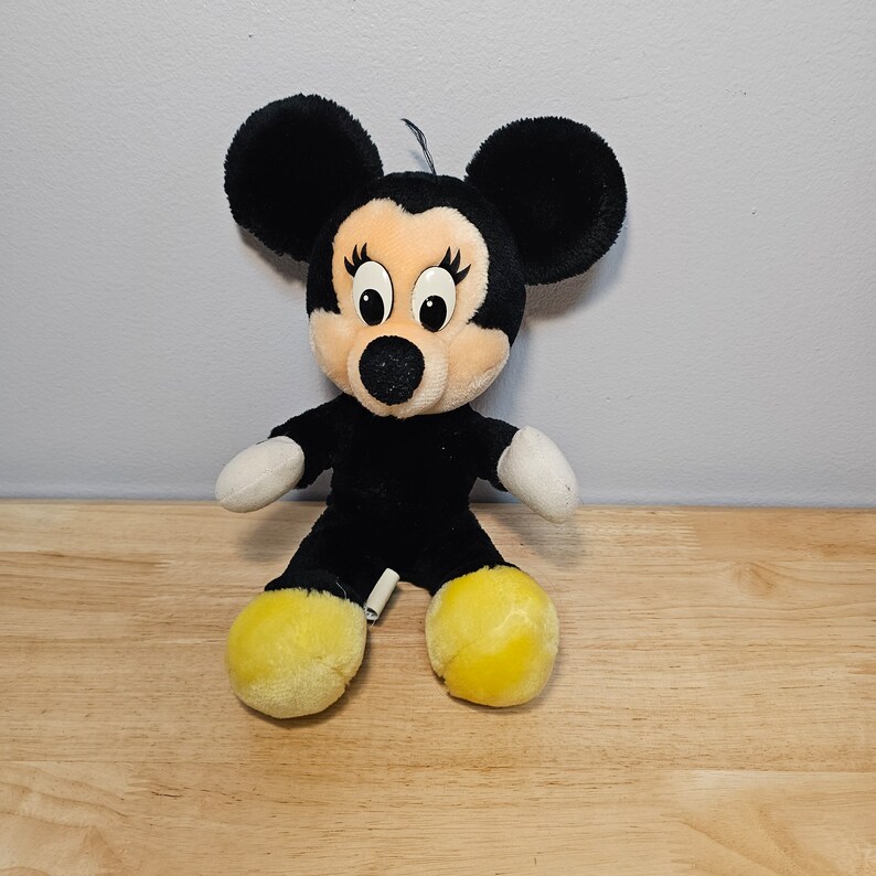 May include: A black and yellow plush toy of Mickey Mouse's girlfriend Minnie Mouse. She has large black ears, a black nose, and yellow shoes.