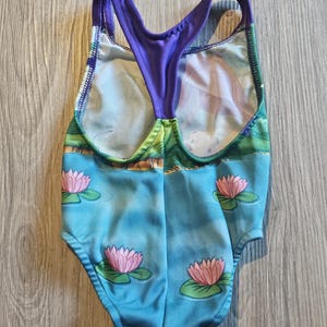 May include: A one-piece swimsuit featuring a blue water scene with pink lotus flowers and green lily pads. The swimsuit has a purple racerback design and a colorful landscape print.