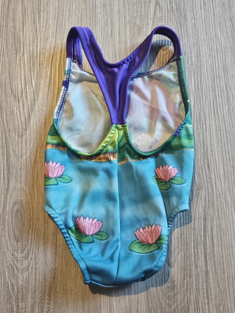 May include: A one-piece swimsuit featuring a blue water scene with pink lotus flowers and green lily pads. The swimsuit has a purple racerback design and a colorful landscape print.