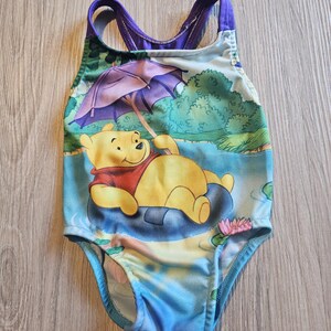 May include: A one-piece swimsuit featuring a cartoon image of Winnie the Pooh relaxing on a lily pad under an umbrella. The swimsuit has a blue, green, and purple color scheme, with a scoop back and straps.