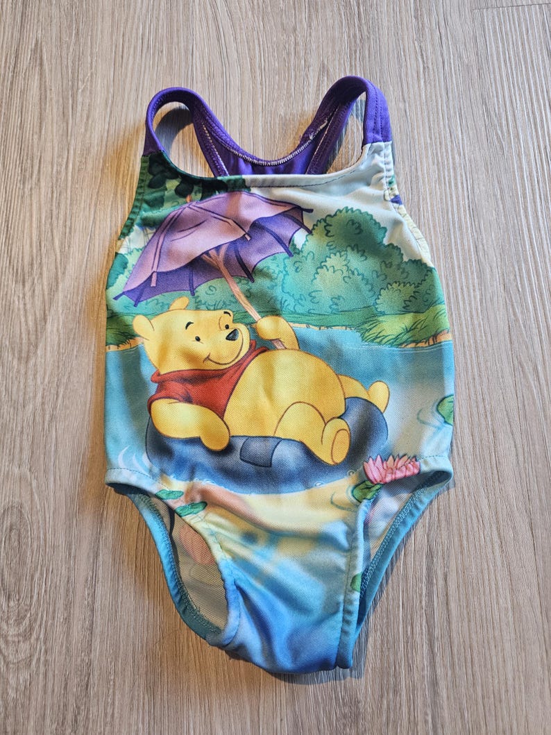 May include: A one-piece swimsuit featuring a cartoon image of Winnie the Pooh relaxing on a lily pad under an umbrella. The swimsuit has a blue, green, and purple color scheme, with a scoop back and straps.