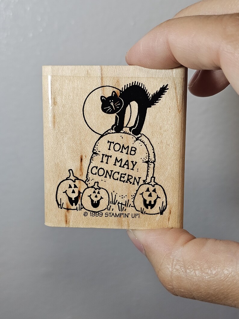 Pode incluir: Um carimbo de madeira com um gato preto em uma l&aacute;pide com a frase "TOMB IT MAY CONCERN" e tr&ecirc;s ab&oacute;boras esculpidas. O carimbo tamb&eacute;m inclui o texto "&copy; 1999 STAMPIN' UP!".