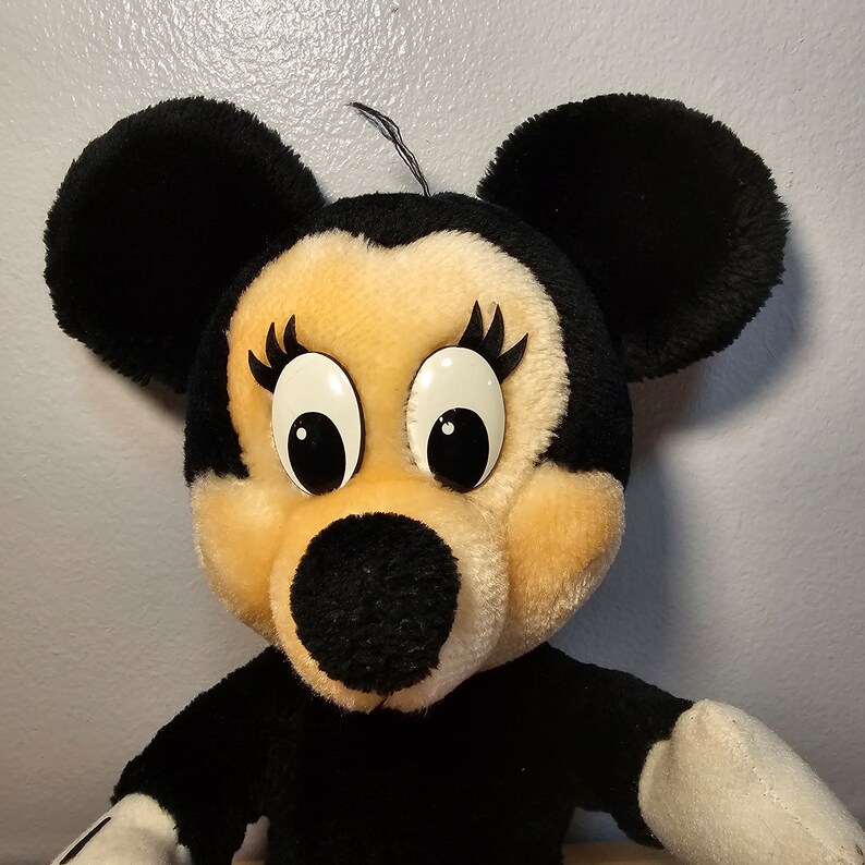 May include: A plush black and white Mickey Mouse character with large eyes and a black nose. The character is wearing a black dress and has white gloves.