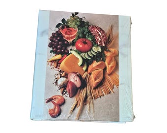 May include: A cookbook with a colorful image of food, including grapes, tomatoes, cheese, bread, and shrimp. The cover has a light blue and white marble design on the edges. The book is wrapped in clear plastic.