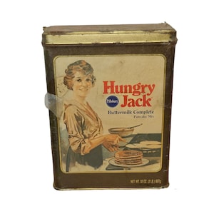 May include: A vintage tin box for Hungry Jack Buttermilk Complete Pancake Mix. The box features a woman in a brown dress holding a spatula over a pan of pancakes. The box is brown with gold trim and has the Pillsbury logo.