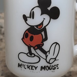 May include: White ceramic mug with a black and red image of Mickey Mouse. The mug has a handle and a base. The text "Mickey Mouse" is printed on the bottom of the mug.