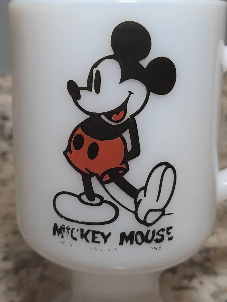 May include: White ceramic mug with a black and red image of Mickey Mouse. The mug has a handle and a base. The text "Mickey Mouse" is printed on the bottom of the mug.