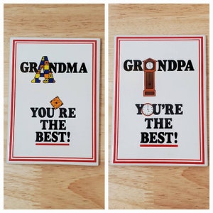 May include: Two white ceramic tiles with red borders. The tile on the left has a colorful patchwork letter A and the text "Grandma You're The Best!" The tile on the right has a brown grandfather clock and the text "Grandpa You're The Best!"