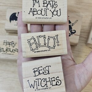 Pode incluir: Carimbos de madeira com frases tem&aacute;ticas de Halloween: "I'm Bats About You", "Boo!" com letras em forma de osso e "Best Witches". Os carimbos est&atilde;o em uma m&atilde;o, com uma silhueta de morcego e "Happy Halloween" vis&iacute;veis.