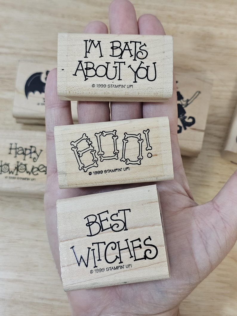 Pode incluir: Carimbos de madeira com frases tem&aacute;ticas de Halloween: "I'm Bats About You", "Boo!" com letras em forma de osso e "Best Witches". Os carimbos est&atilde;o em uma m&atilde;o, com uma silhueta de morcego e "Happy Halloween" vis&iacute;veis.