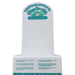 May include: A white display stand with a teal arch at the top that reads "applause presents Cabbage Patch Kids Porcelain Dolls". The stand has text describing the dolls as limited edition and created by Xavier Roberts. The text also mentions that the dolls are made from fine porcelain china and each doll wears exquisite handmade clothing. The stand has the words "LIMITED EDITION" at the bottom.