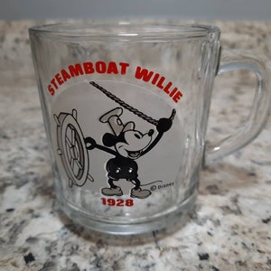 May include: A clear glass mug with a black and white image of Mickey Mouse as Steamboat Willie. The mug has the text "Steamboat Willie" in red lettering and "1928" in black lettering.