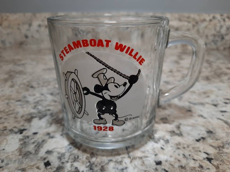 May include: A clear glass mug with a black and white image of Mickey Mouse as Steamboat Willie. The mug has the text "Steamboat Willie" in red lettering and "1928" in black lettering.