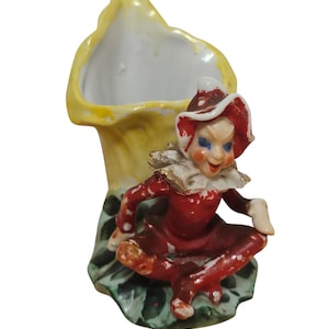 May include: A vintage ceramic figurine featuring a seated elf in a red outfit with a white collar and hat. The elf is positioned in front of a yellow flower-shaped vase with a green base. The figurine has blue eyes and a whimsical expression.
