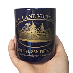 May include: A blue ceramic mug with gold lettering that reads "S.S. LANE VICTORY" and "BERTH 94, SAN PEDRO, CA. National Historic Landmark". The mug features a gold illustration of a ship.