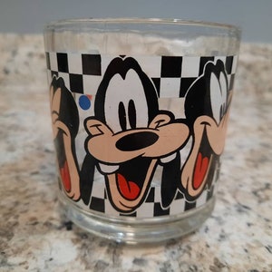 May include: A clear glass tumbler with a black and white checkered pattern and three images of Goofy. Goofy is depicted with a black nose, red mouth, and white eyes.