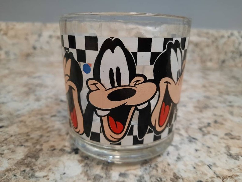 May include: A clear glass tumbler with a black and white checkered pattern and three images of Goofy. Goofy is depicted with a black nose, red mouth, and white eyes.