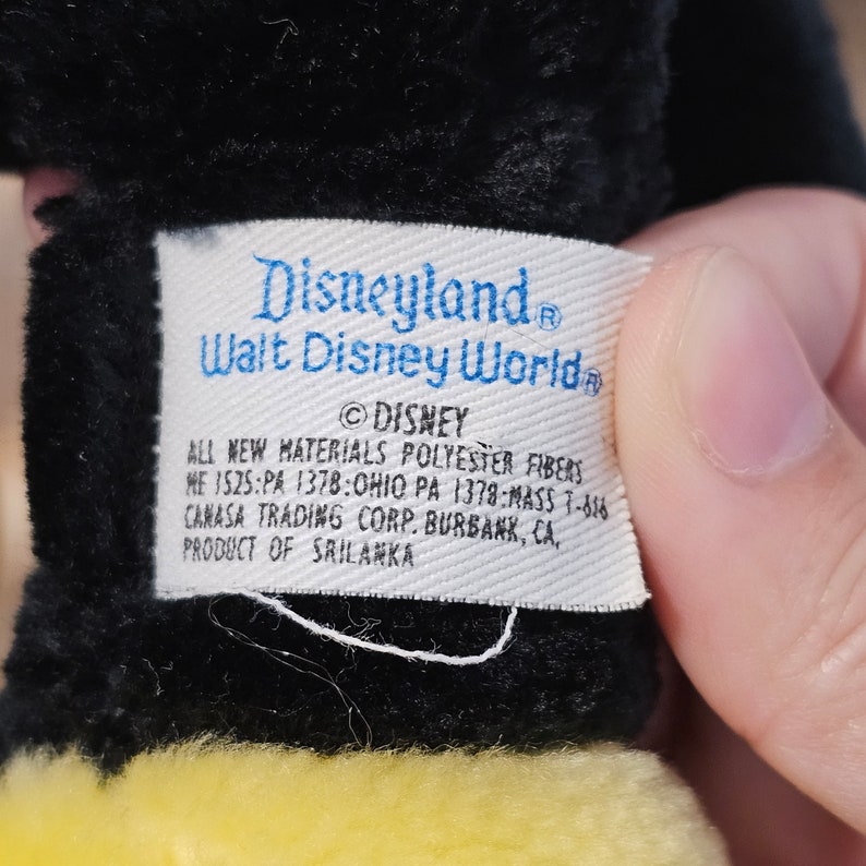 May include: A white tag with blue text that reads "Disneyland Walt Disney World &copy;DISNEY ALL NEW MATERIALS POLYESTER FIBERS ME ISZS:PA 1378:OHIO PA 1378:MASS 7-616 CANASA TRADING CORP. BURBANK, CA, PRODUCT OF SRILANKA"