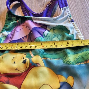 May include: A children's swimsuit featuring Winnie the Pooh. The one-piece swimsuit has a purple and green design with a yellow measuring tape showing a length of approximately 8 inches. The swimsuit has a scoop back and straps.