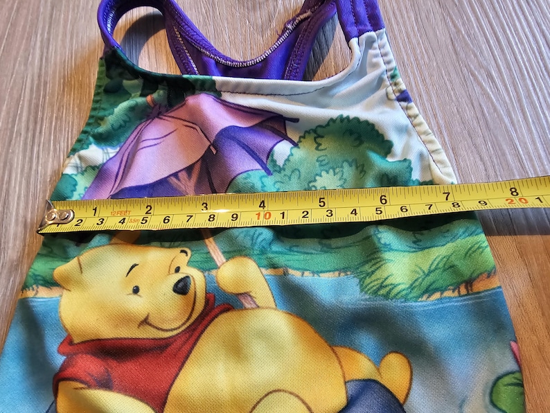 May include: A children's swimsuit featuring Winnie the Pooh. The one-piece swimsuit has a purple and green design with a yellow measuring tape showing a length of approximately 8 inches. The swimsuit has a scoop back and straps.
