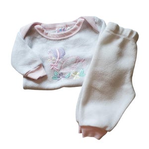May include: A white and pink baby outfit with a long-sleeved shirt and pants. The shirt features an embroidered design of a duck with flowers.