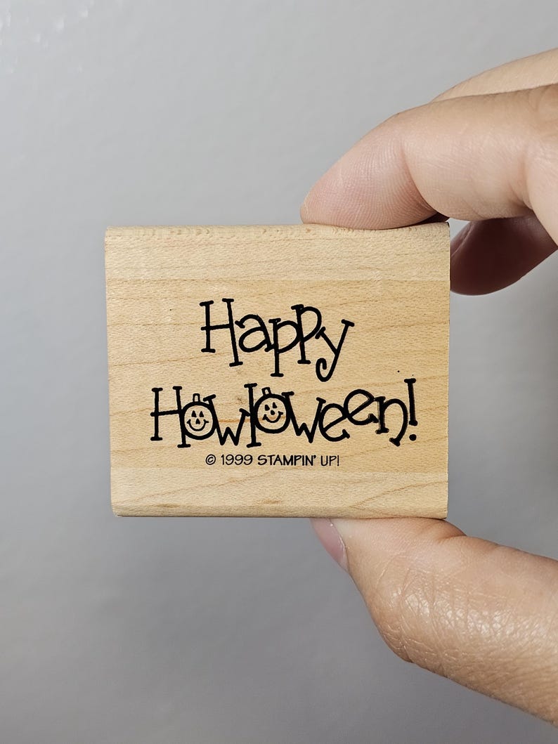 Pode incluir: Um carimbo de madeira com as palavras "Happy Halloween!" em preto, com uma fonte extravagante. A palavra "Halloween" apresenta rostos de jack-o'-lantern em vez das letras "o". O carimbo tamb&eacute;m inclui o texto "&copy; 1999 STAMPIN' UP!"