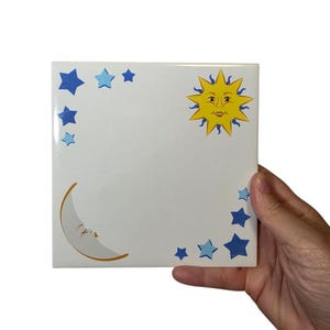 May include: A square, white ceramic tile featuring a celestial design. The tile has a yellow sun with a smiling face, a crescent moon, and blue stars. The tile is being held by a hand.
