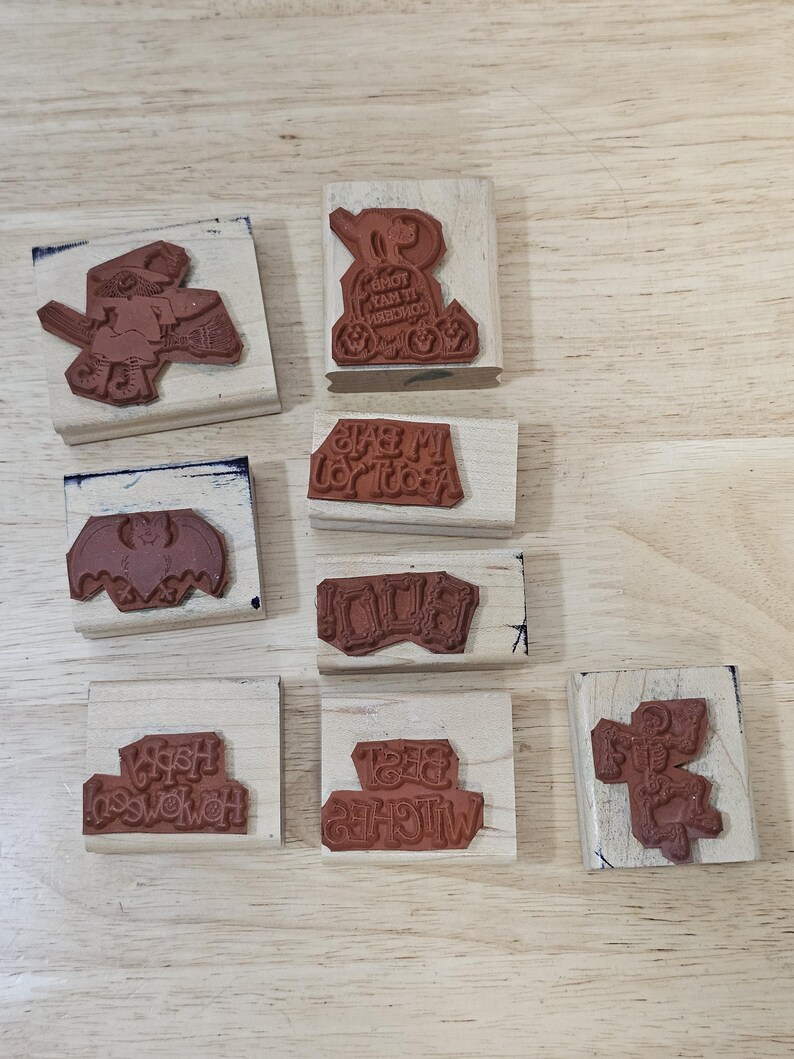 Pode incluir: Uma cole&ccedil;&atilde;o de carimbos de borracha castanhos em blocos de madeira. Os carimbos apresentam desenhos tem&aacute;ticos de Halloween, incluindo uma bruxa, um morcego e textos como "Happy Halloween" e "Best Witches".