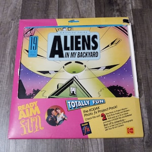 May include: A pink and yellow box with a picture of a flying saucer over a house. The box is labeled "Gripping Tales" and "Aliens in My Backyard". The box also has a picture of two people holding a camera and the text "Ready Aim Fun!" and "Totally Fun! The Kodak Photo fx Project Pack!"