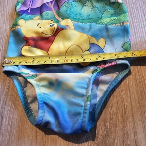 May include: A baby swimsuit featuring a Winnie the Pooh design. The swimsuit is primarily blue with a colorful print of the character. The measurement across the top is approximately 9 inches.