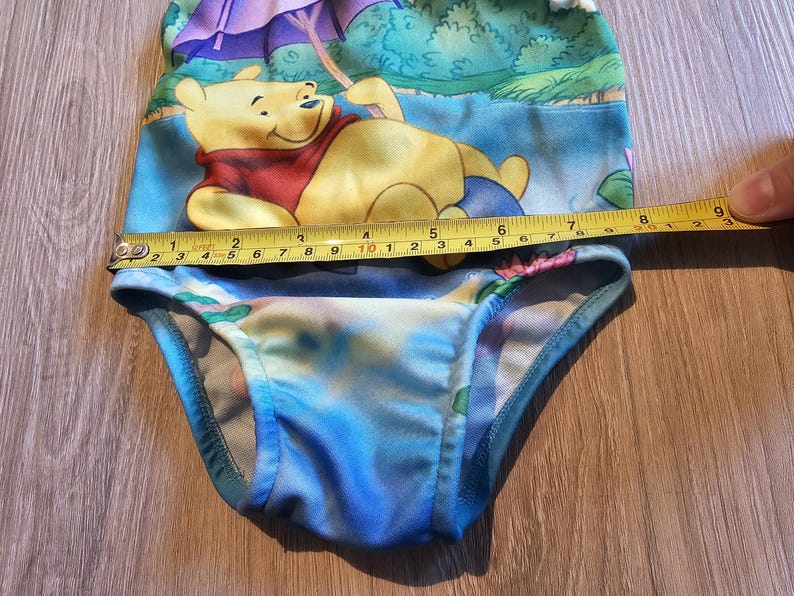 May include: A baby swimsuit featuring a Winnie the Pooh design. The swimsuit is primarily blue with a colorful print of the character. The measurement across the top is approximately 9 inches.