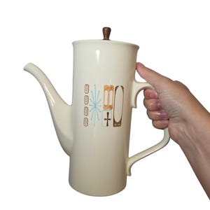 May include: A tall, cream-colored ceramic coffee pot featuring a mid-century modern graphic pattern in light blue and brown. The design includes vertical lines, a starburst, and rectangular shapes. It has a long spout, a handle, and a brown lid knob.