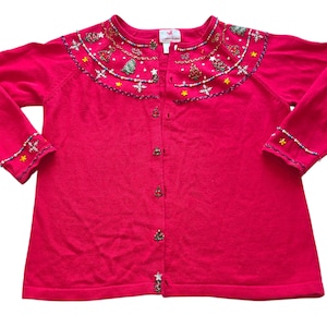 May include: A bright pink cardigan sweater with long sleeves. The collar and cuffs are embellished with beads and small Christmas-themed designs. The sweater has a button-up front and is a festive holiday garment.