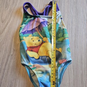 May include: A one-piece swimsuit featuring Winnie the Pooh. The swimsuit has a colorful design with a yellow Winnie the Pooh character, a purple umbrella, and a blue and green background. The swimsuit measures approximately 13 inches in length.