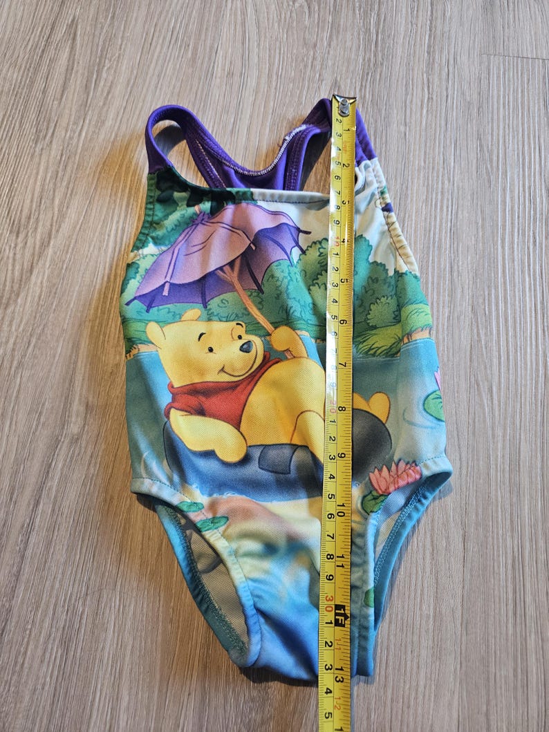 May include: A one-piece swimsuit featuring Winnie the Pooh. The swimsuit has a colorful design with a yellow Winnie the Pooh character, a purple umbrella, and a blue and green background. The swimsuit measures approximately 13 inches in length.