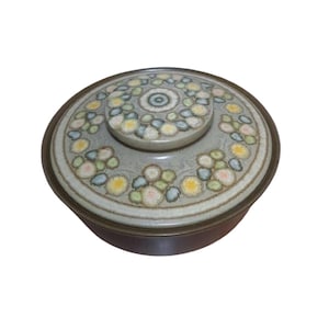 May include: A round ceramic serving dish with a lid. The dish has a brown rim and a white base with a floral pattern in shades of green, blue, yellow, and pink.