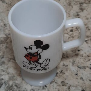May include: White ceramic mug with a black and white image of Mickey Mouse wearing red shorts. The mug has a pedestal base and says "Mickey Mouse" on the bottom.