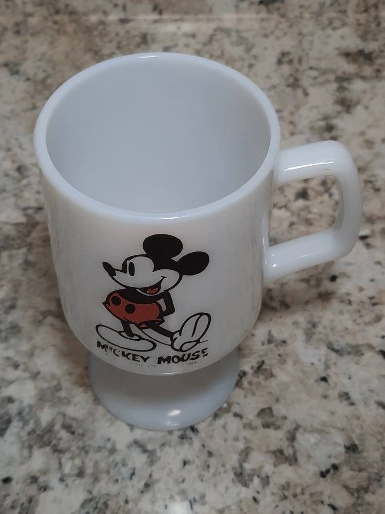 May include: White ceramic mug with a black and white image of Mickey Mouse wearing red shorts. The mug has a pedestal base and says "Mickey Mouse" on the bottom.