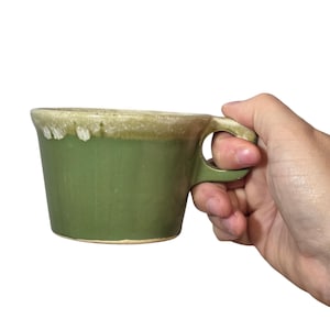 May include: A green ceramic mug with a rounded handle. The mug has a slightly flared shape and a light green rim. The mug is held by a hand against a white background. The mug is likely handmade.