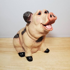 May include: A ceramic pig figurine with black and brown markings. The pig is sitting with its mouth open and has a happy expression.
