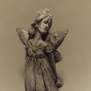 Może przedstawiać: Szara kamienna figurka wróżki z skrzydłami, sukienką i kwiatem w ręku. Wróżka stoi na podstawie.