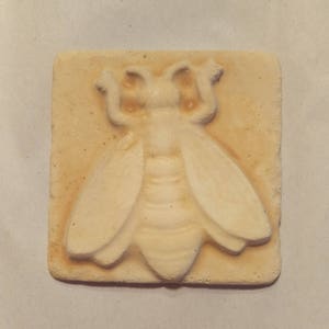May include: A beige ceramic tile with a raised design of a bee. The bee is white and has a textured surface.