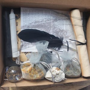 May include: An open cardboard box filled with various items. Includes a black feather, a clear crystal ball, small mesh bags with dried herbs, a rolled parchment, a white candle, and printed papers. The items are arranged inside the box.