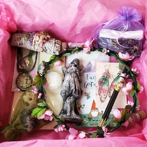 May include: A pink gift box filled with various items, including a small stone angel figurine, a floral wreath, a sage smudge stick, a small bottle, and a card that says "Fairy Garden".