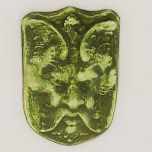 May include: A green ceramic tile featuring a raised design of a grotesque face with wings. The face is surrounded by floral details.