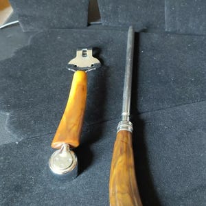 May include: A kitchen tool set consisting of a metal peeler and a sharpening steel, both featuring curved, amber-coloured handles with a marbled pattern. The peeler has a silver-toned metal head, and the sharpening steel has a long, slender metal rod.