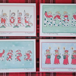May include: Four Christmas cards with watercolor illustrations. Each card features the word "NOEL" or a variation, with festive imagery. One card shows children with candles, another has angels, and two others have elves. The cards are on a red and green plaid background.