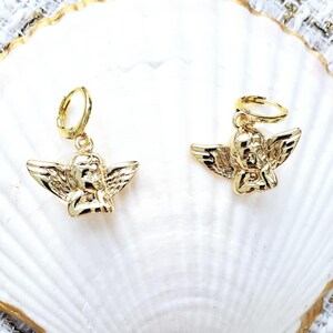 May include: A pair of gold angel-shaped earrings with hoop closures. The earrings are displayed on a white seashell.