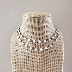 May include: A white and gold layered necklace with small round beads. The necklace is displayed on a burlap mannequin.