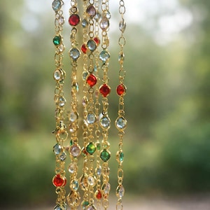May include: A collection of gold-toned chains featuring bezel-set faceted crystals in various colors including red, green, light blue, and clear. The delicate metal links are arranged vertically, showcasing a repeating pattern of colorful stones.